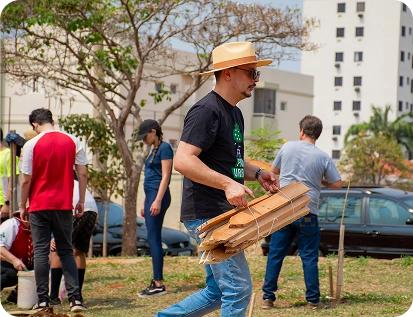 oluntário de chapéu e camiseta do projeto carrega um fardo de estacas de madeira em meio a um canteiro urbano, preparando a estrutura de suporte para as novas mudas que serão plantadas pela equipe.